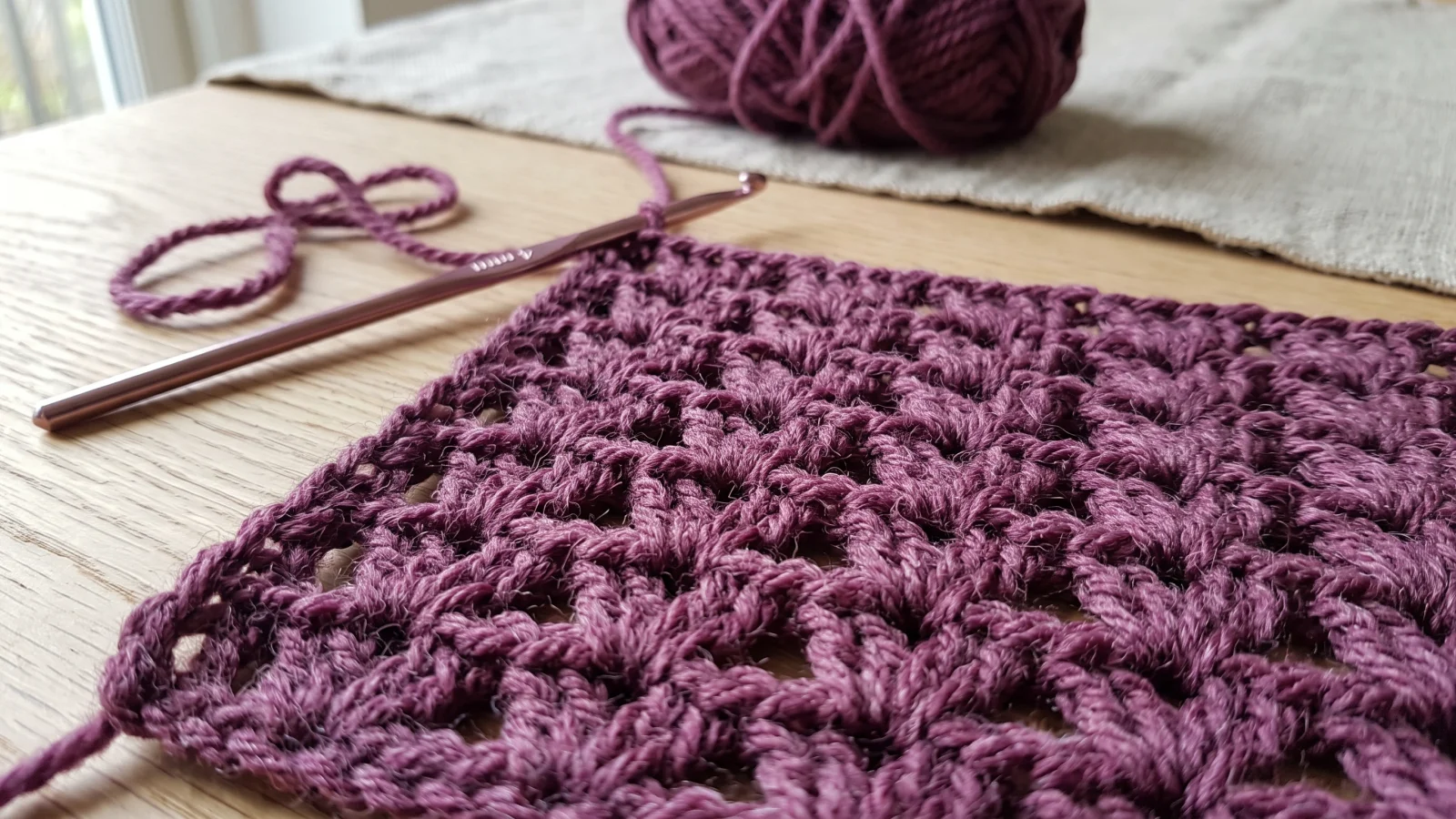 Close-up of plum crochet V-stitch fabric showing rows of paired half double crochets with chain-one spaces, beside a crochet hook.